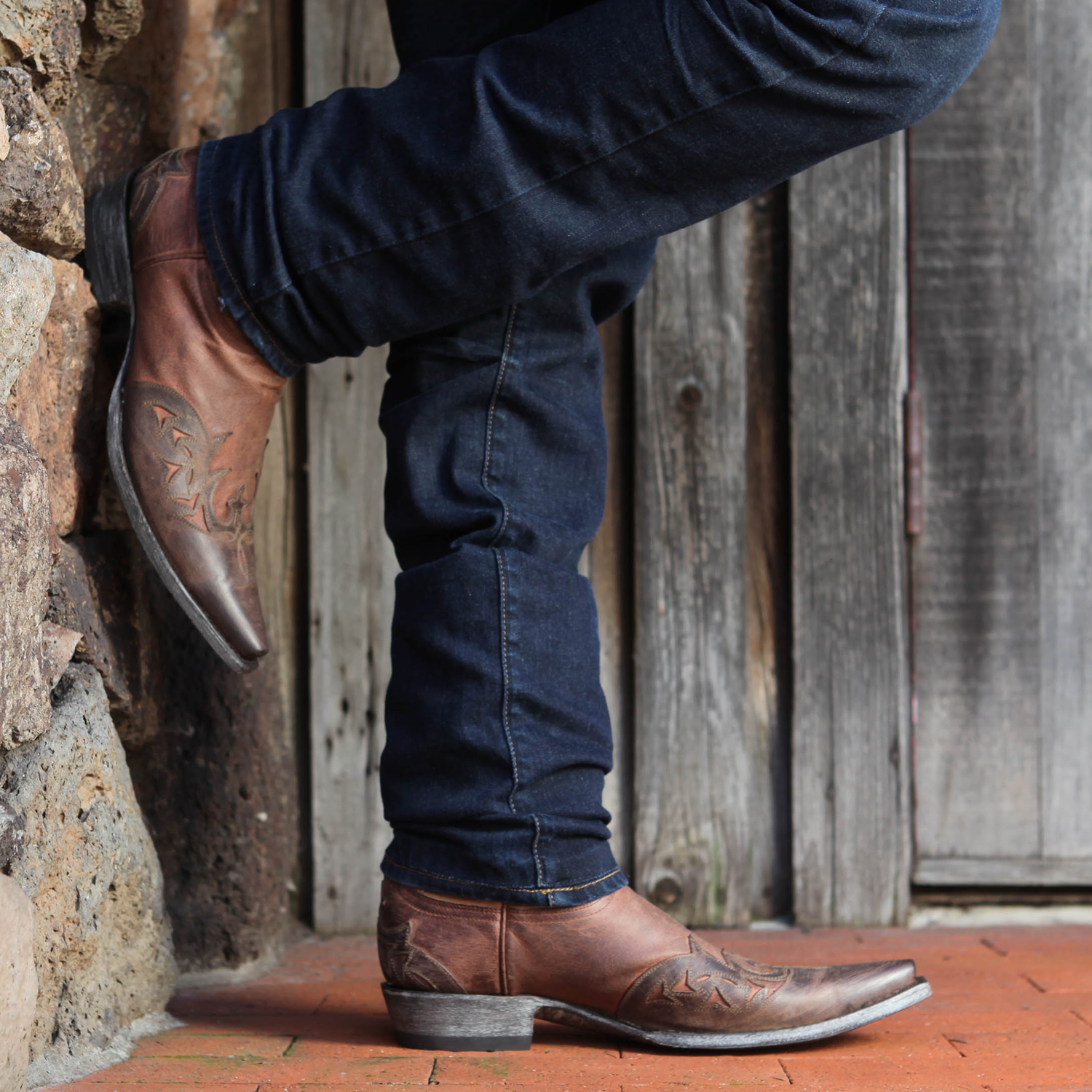 A man in dark blue jeans wearing brown boots with a cactus design in a rustic background